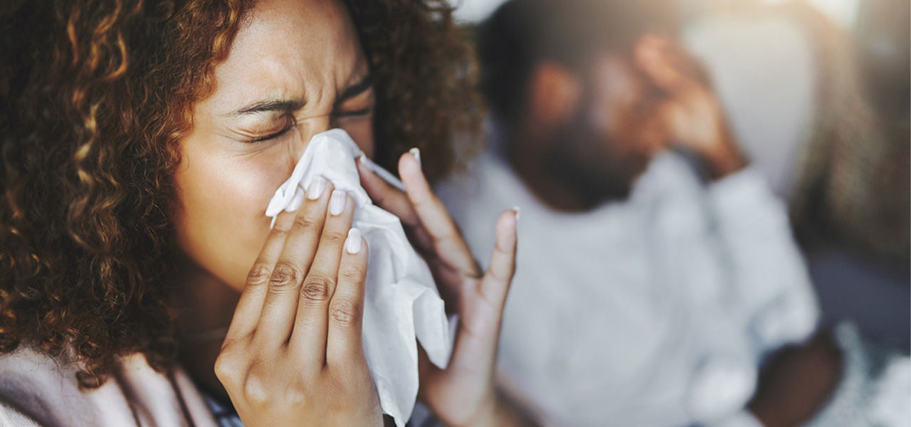 woman sneezing into tissue next to ailing husband with headache and flu symptoms
